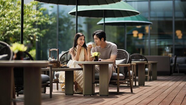 Una pareja cenando en un café al aire libre con sombrillas verdes y muebles de mimbre.