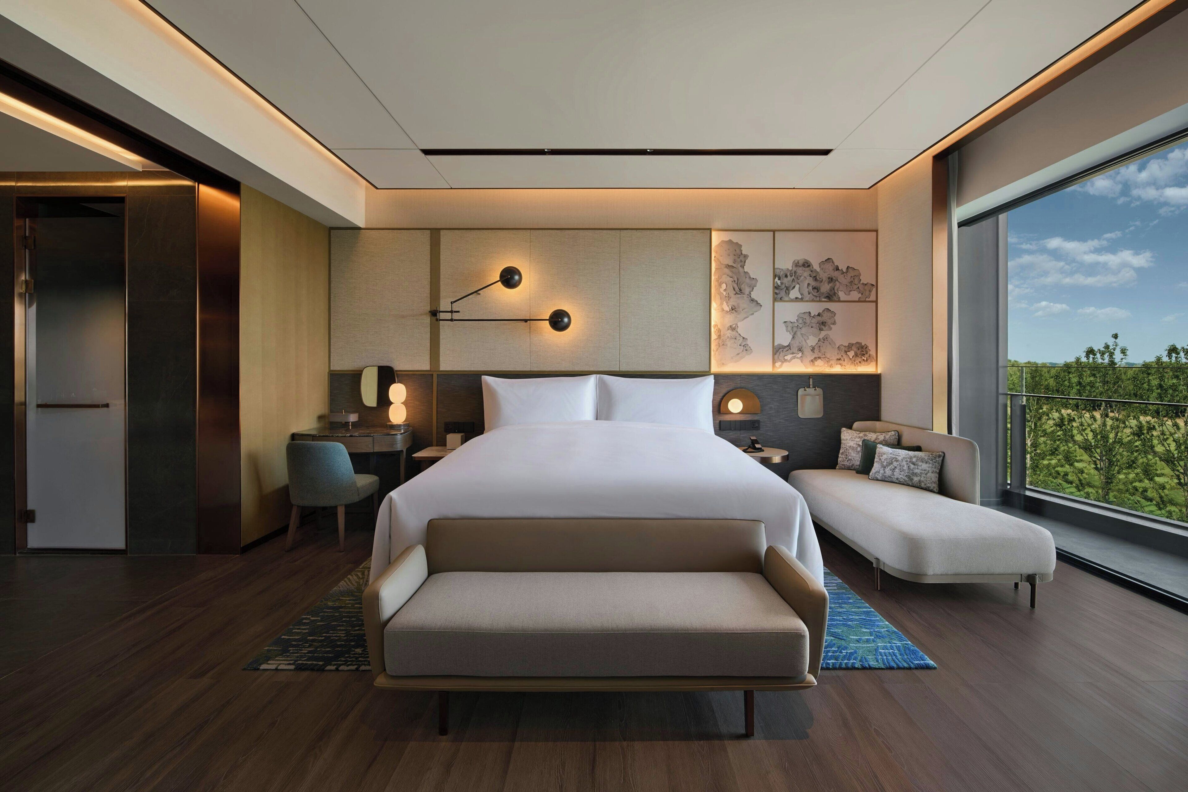 Un dormitorio moderno con una cama grande, un sofá, un escritorio y vistas a zonas verdes por la ventana.
