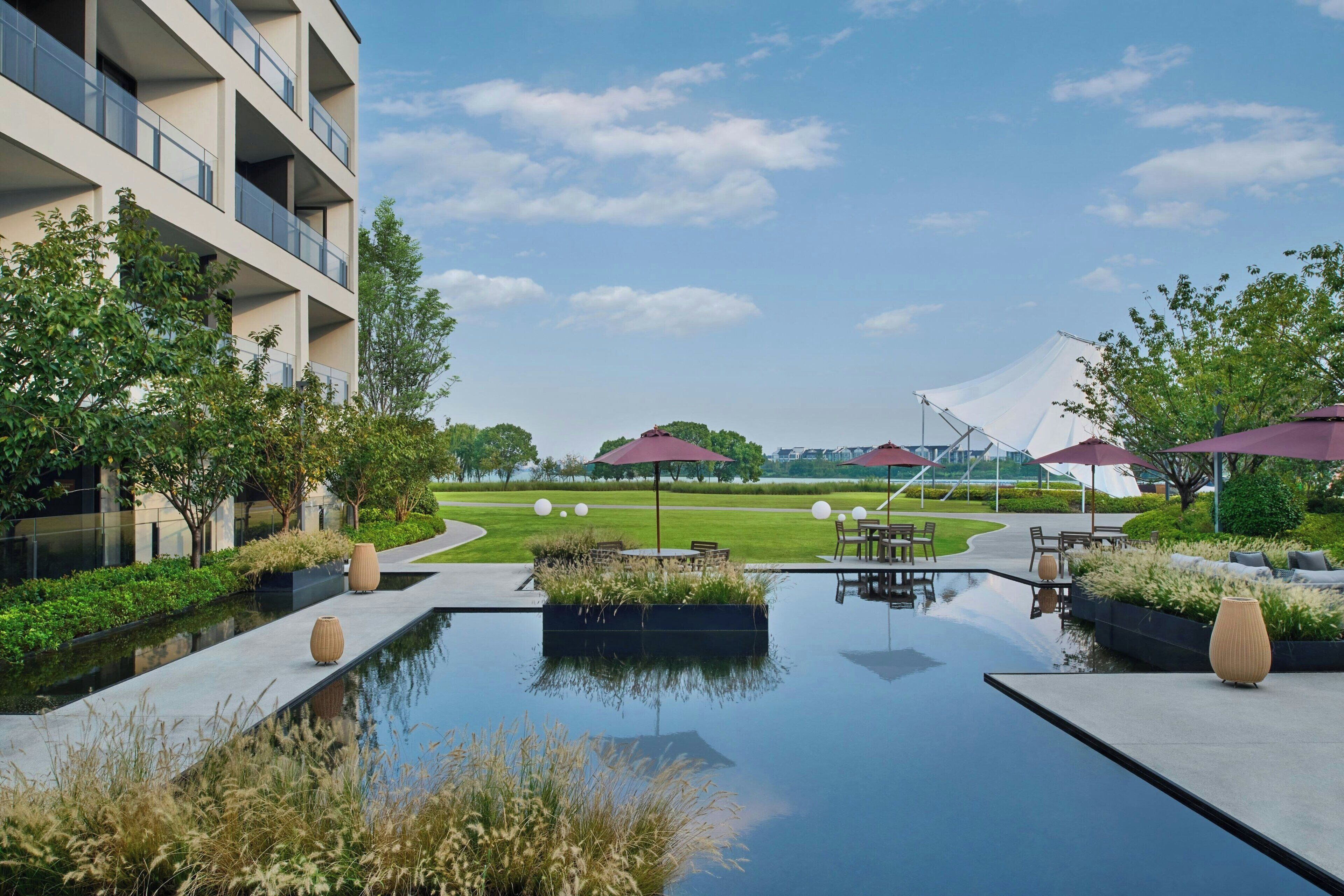 A modern outdoor area with a reflective pool, seating areas, and a building with balconies.