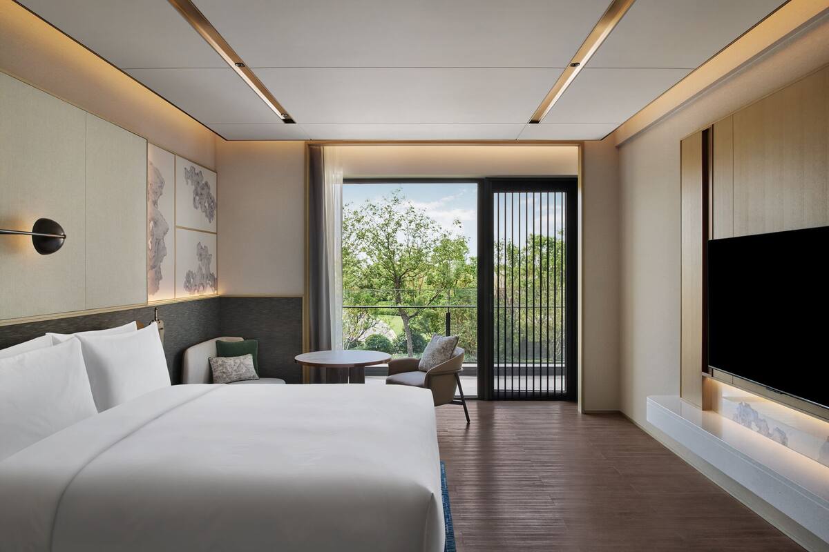 Ein modernes Hotelzimmer mit einem großen Bett, einem Fernseher, einem Balkon mit Blick auf Bäume und einem kleinen Tisch mit Stühlen.
