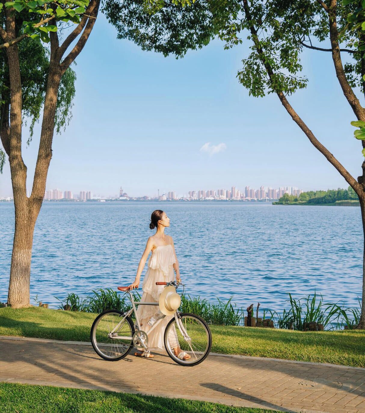 A person walking a bicycle along a paved path beside a body of water with a city skyline in the background.
