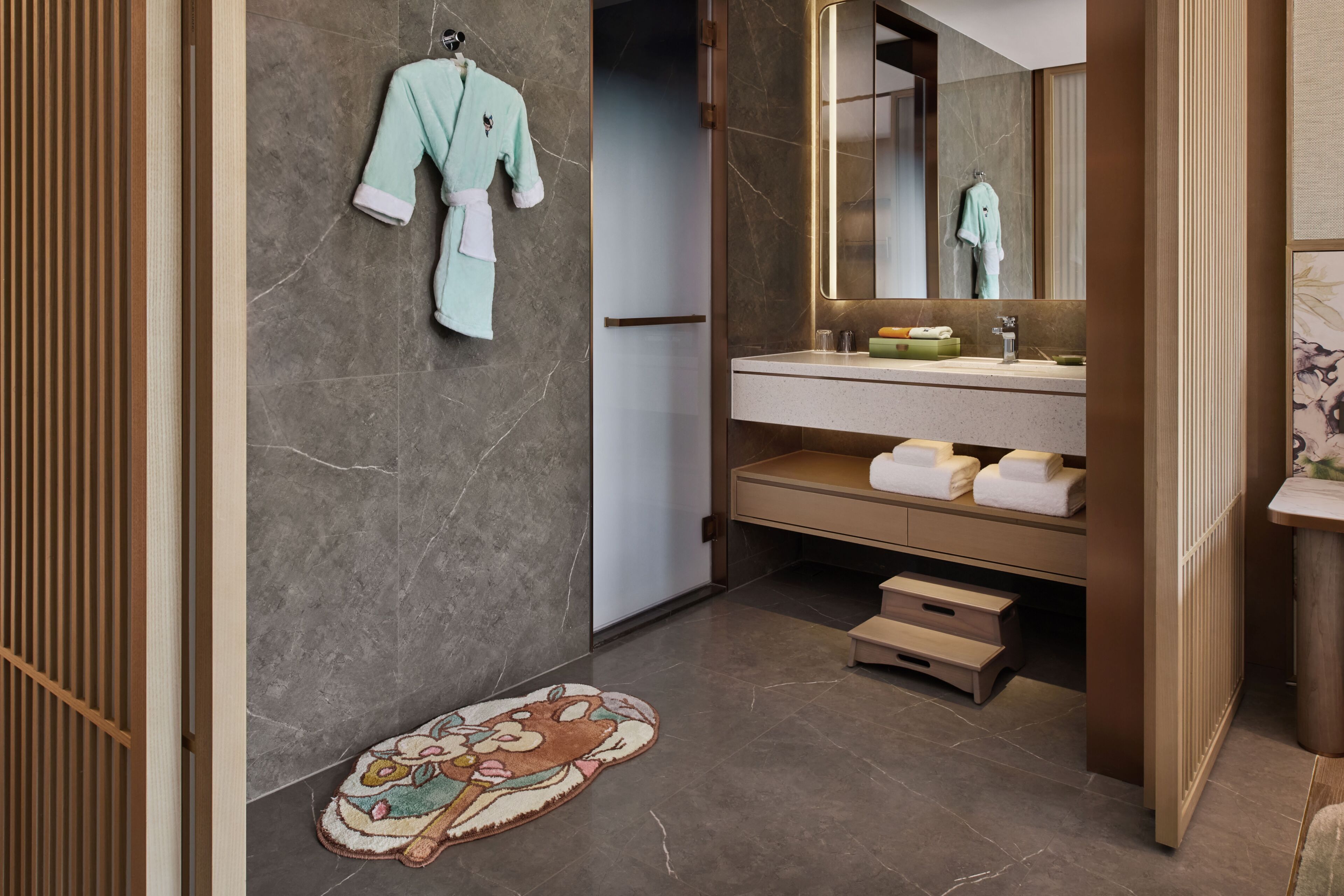 Un baño con pared de mármol, lavamanos con toallas, espejo y suelo de madera.