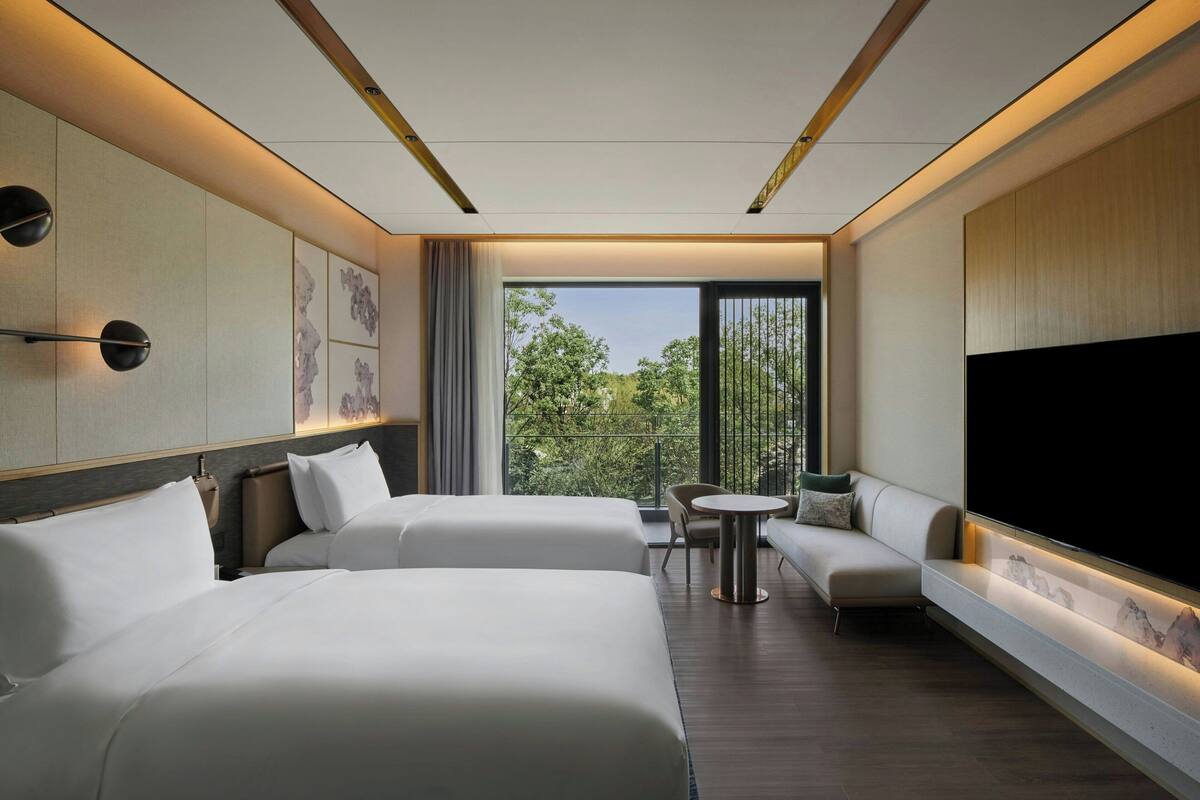 Ein modernes Hotelzimmer mit zwei Betten, einem Sofa, einem Fernseher und einem Balkon mit Blick auf Bäume.