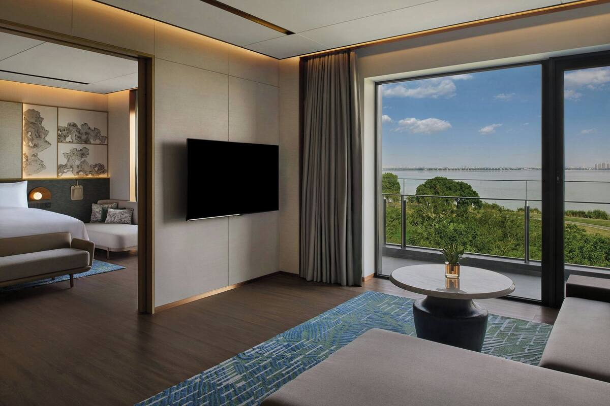 Ein modernes Hotelzimmer mit einem großen Bett, einem Flachbildfernseher, einem Balkon mit Blick auf die Stadt und das Wasser sowie einem kleinen Tisch mit einer Vase.