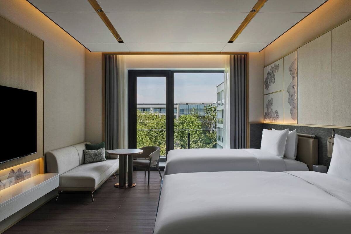 Ein modernes Hotelzimmer mit einem großen Bett, einer Couch, einem kleinen Tisch und einem Fernseher. Das Zimmer verfügt über einen Balkon mit Blick auf Gebäude und Bäume.