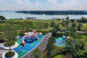 Un parque infantil con toboganes y estructuras para escalar, rodeado de vegetación y vistas a un lago tranquilo y edificios lejanos.
