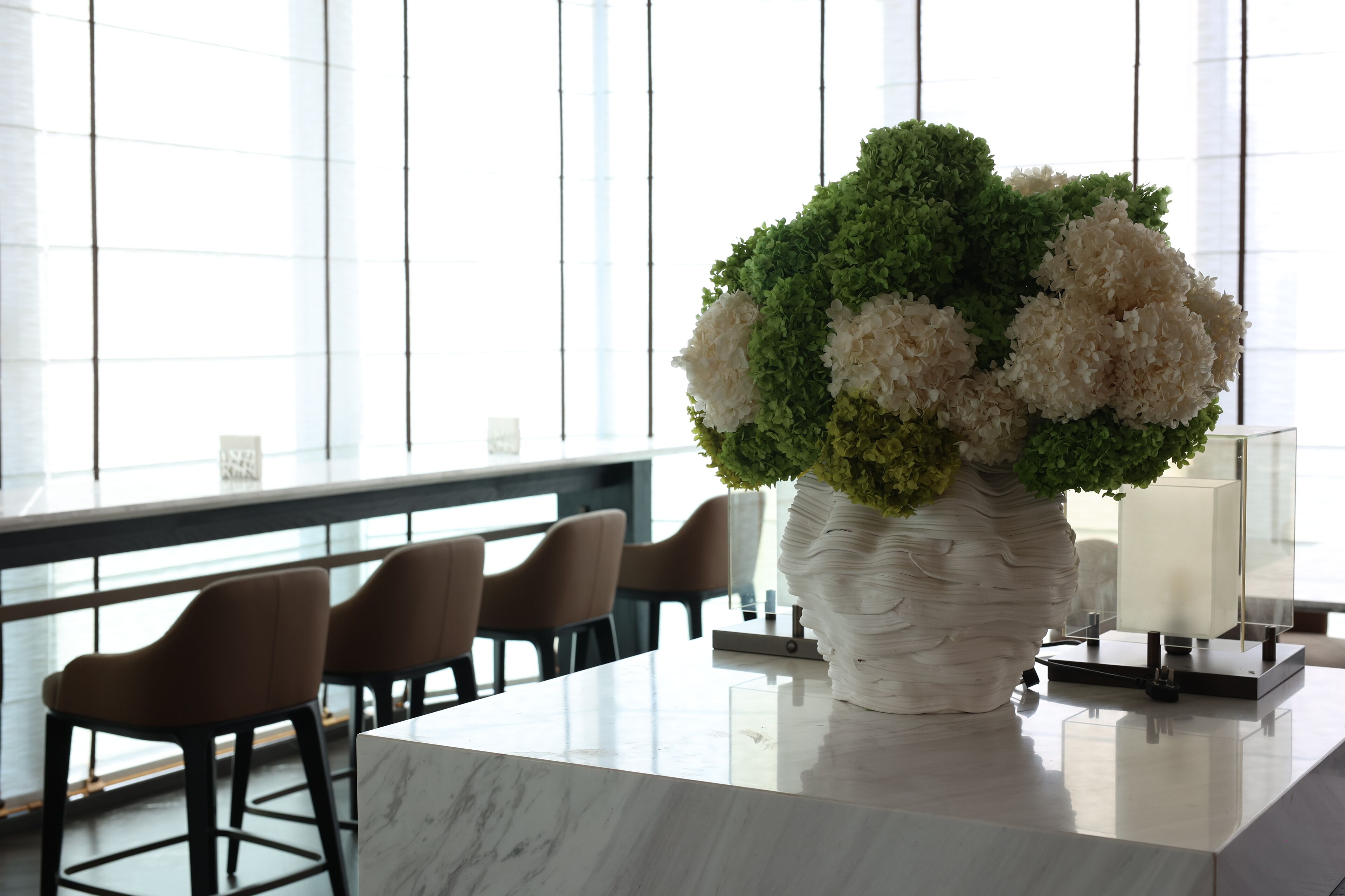 A modern hotel lobby with a marble reception desk, a large potted plant with white flowers, and a seating area with brown chairs.