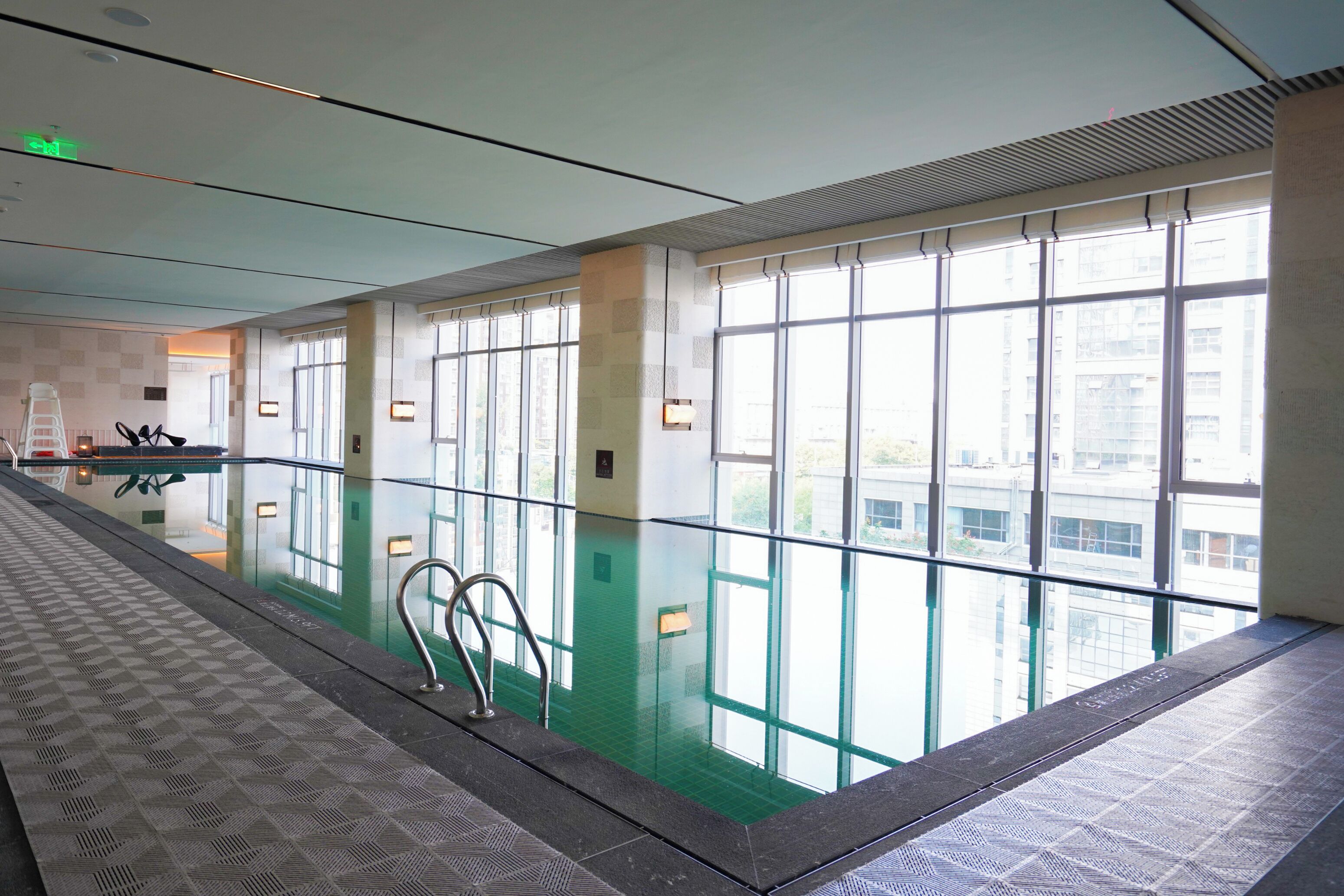 A swimming pool with glass railings, a tiled floor, and a view of buildings through large windows.