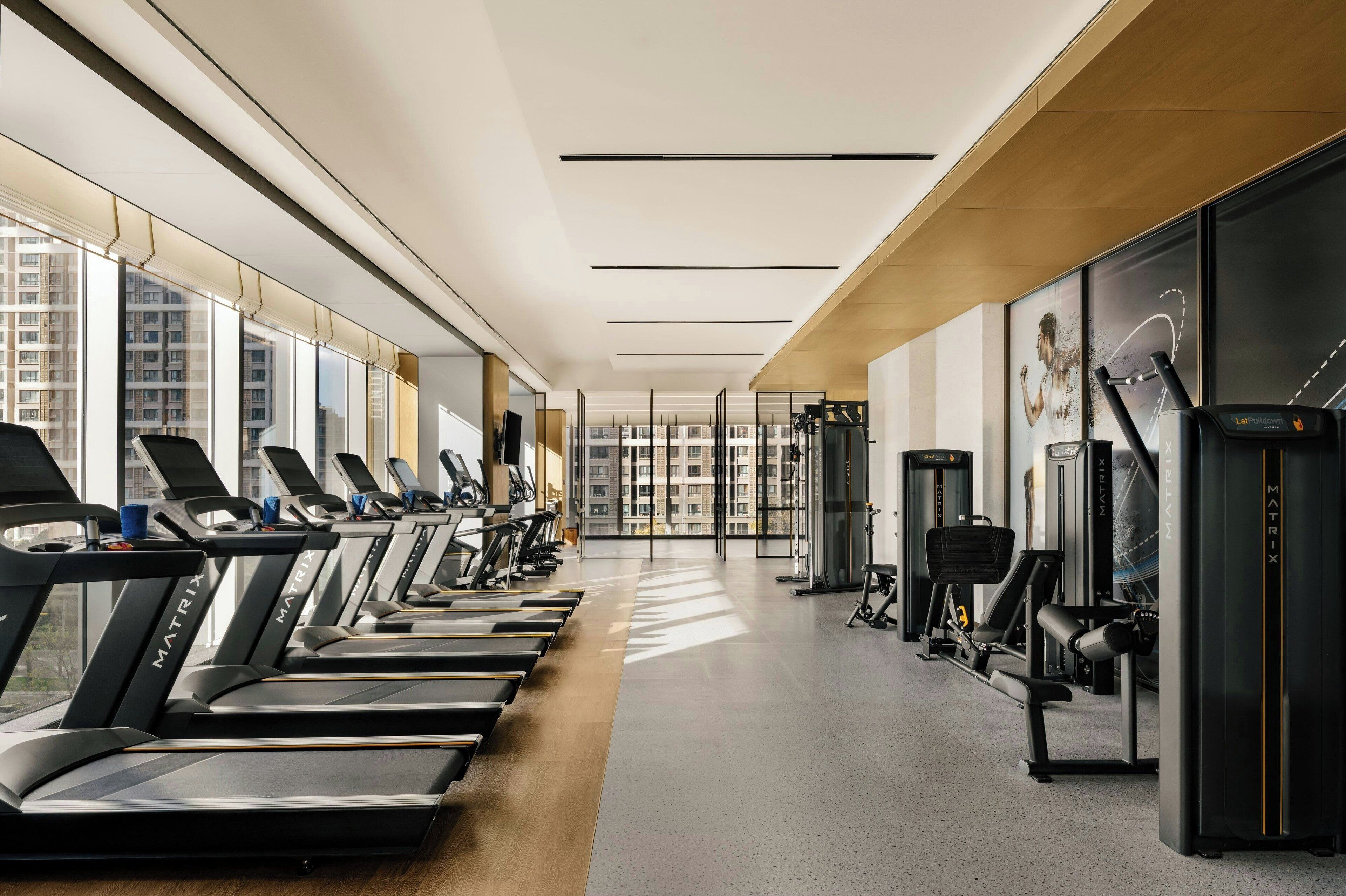 Un gimnasio con cintas de correr, máquinas de ejercicio y amplias ventanas con vistas a edificios.