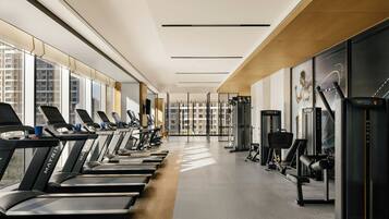Un gimnasio con cintas de correr, máquinas de ejercicio y amplias ventanas con vistas a edificios.