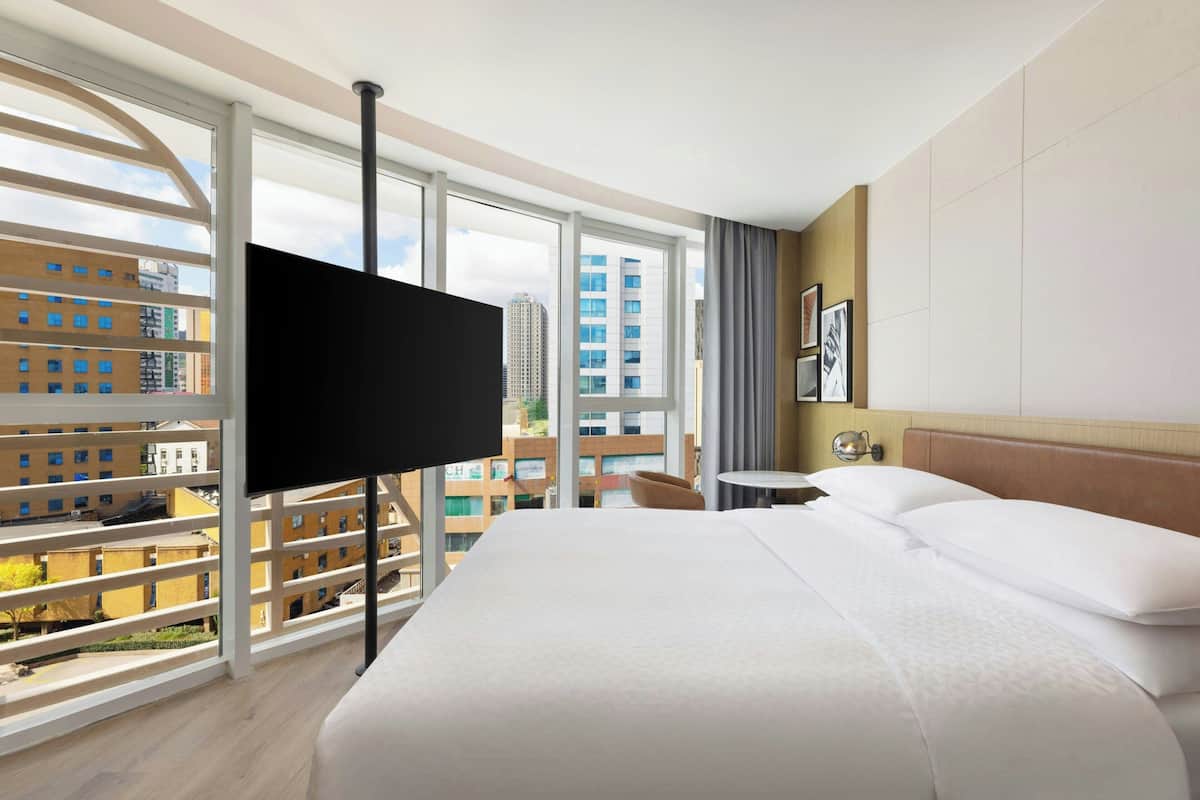 Een hotelkamer met een groot bed, een televisie aan een witte muur bevestigd, een klein tafeltje en uitzicht op gebouwen door grote ramen.