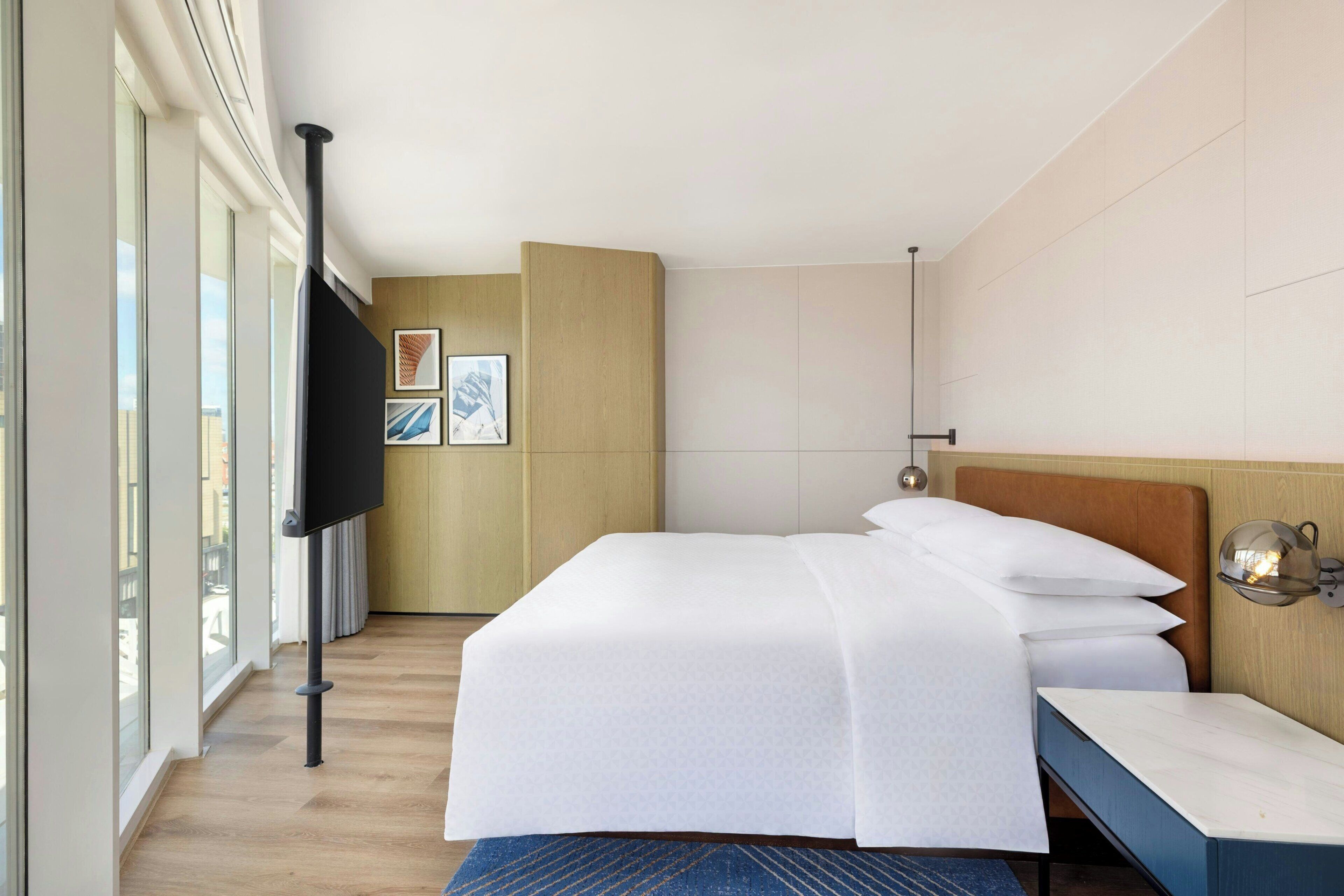 Habitación de hotel moderna con una cama grande, televisión de pantalla plana, cabecero de madera y vistas amplias por grandes ventanas.