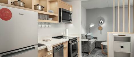 A modern kitchen with a built-in microwave, oven, and dishwasher. There is a wooden cabinet above the counter with pots and a microwave. A small seating area with a sofa and a table is visible in the background.