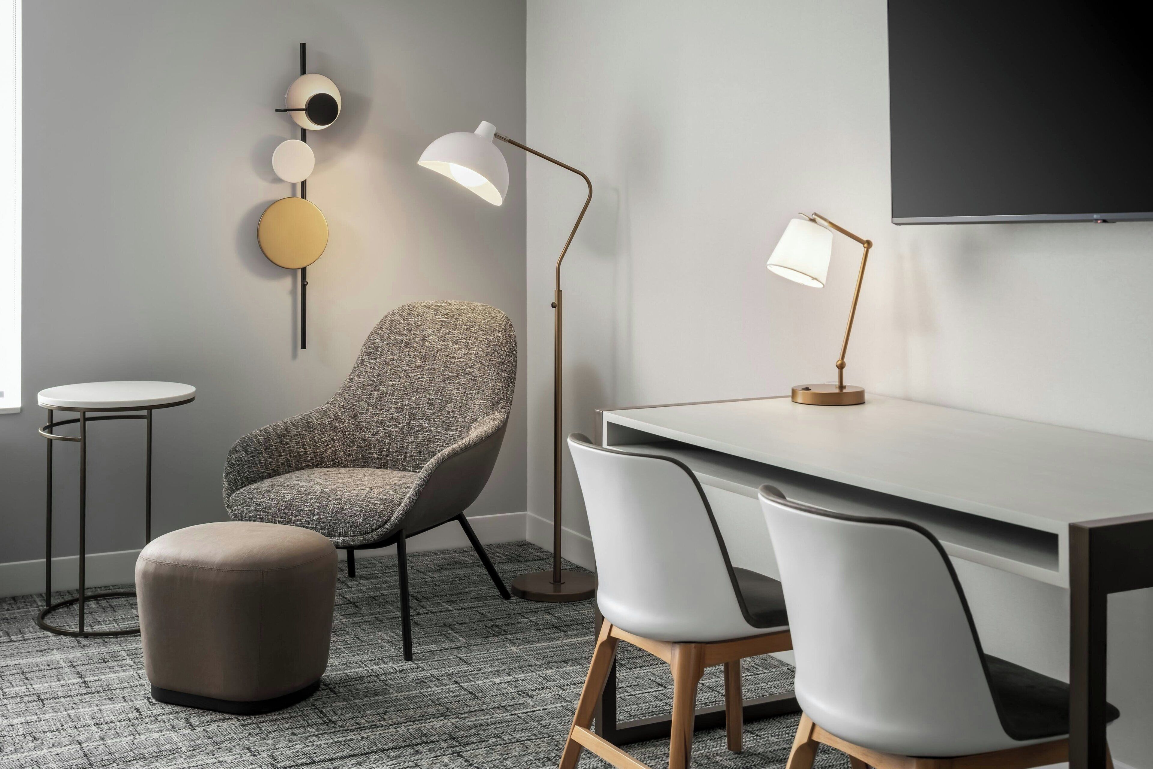 A modern office space with a desk, two chairs, a round ottoman, a floor lamp, a standing lamp, and a wall-mounted spherical object.