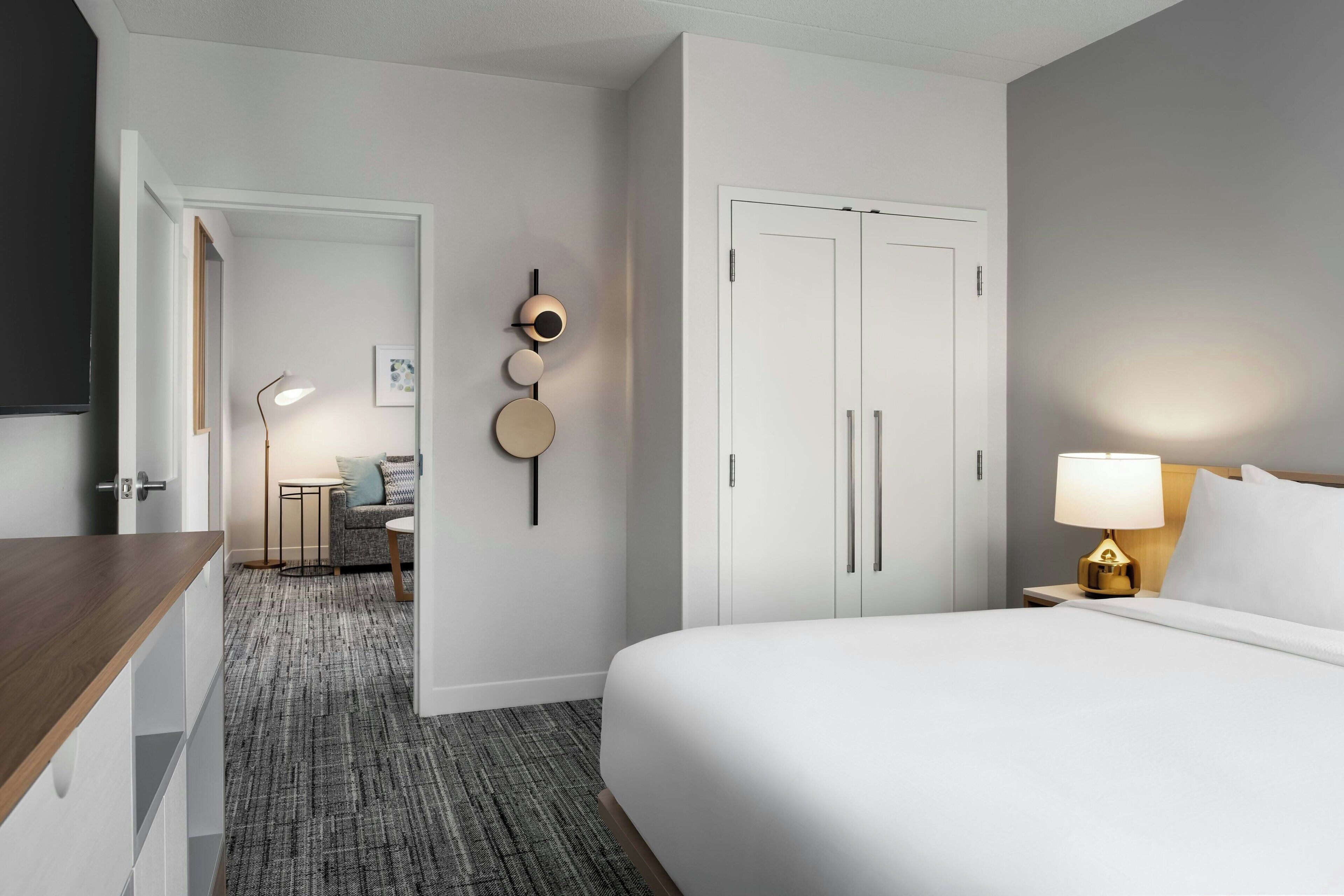 A hotel room with a bed, bedside table, lamp, wardrobe, and a small seating area with a table and chairs.