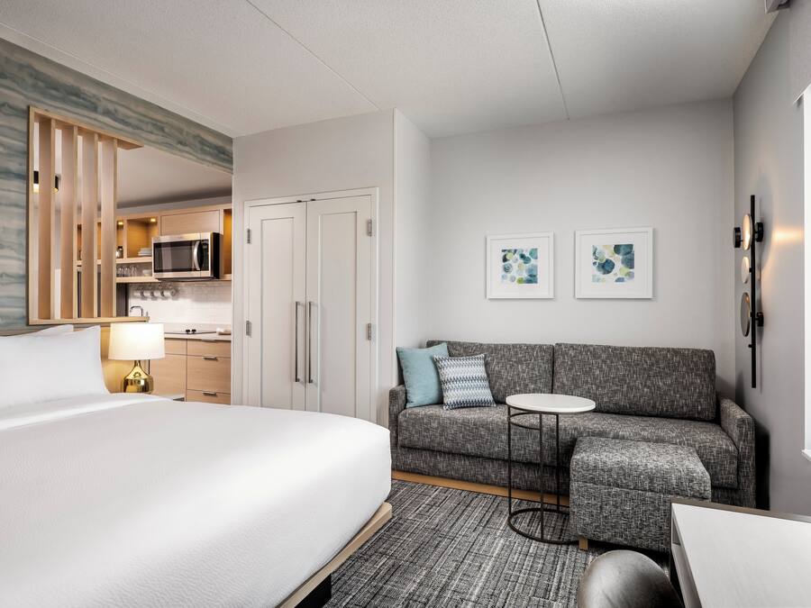 A hotel room with a bed, a gray sofa, a small round table, and a kitchenette in the background.