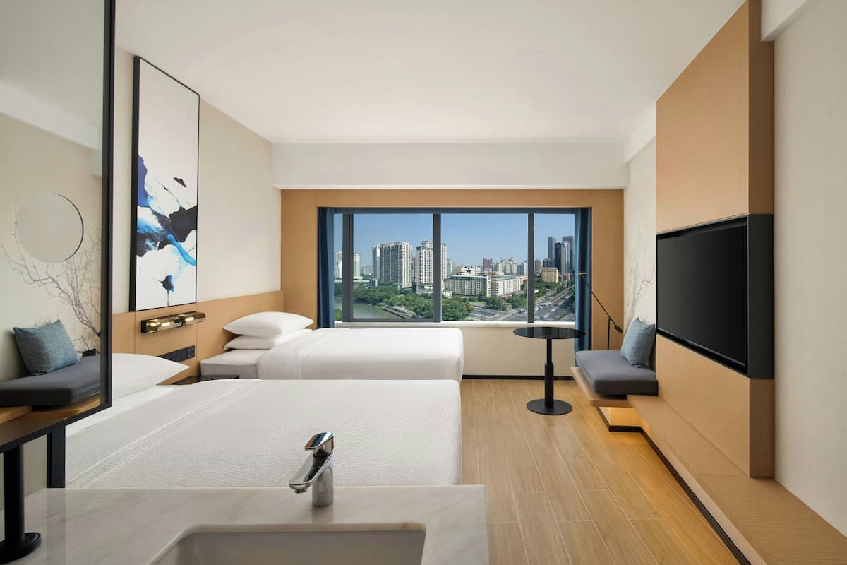 A modern hotel room with a large window offering a city view, a bed, a sofa, a TV, and a bathroom with a sink.
