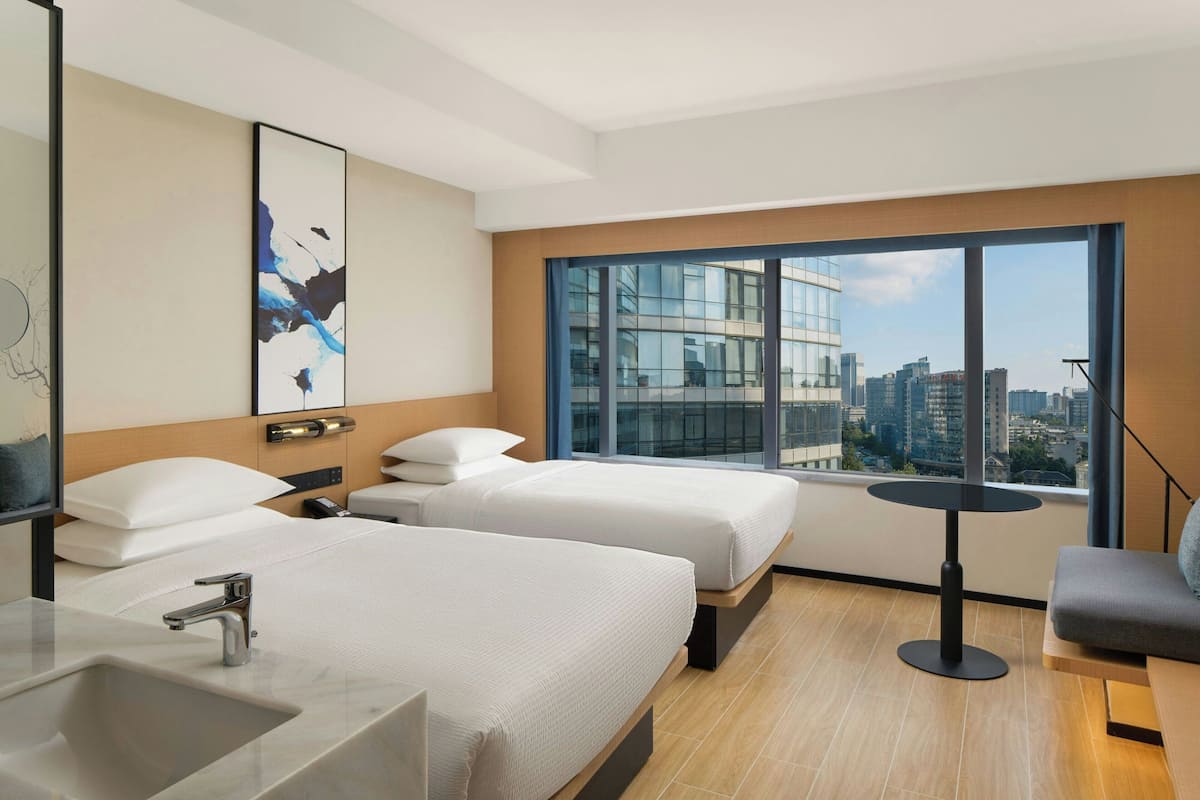 A hotel room with two beds, a large window with a city view, a round table, and a modern bathroom.