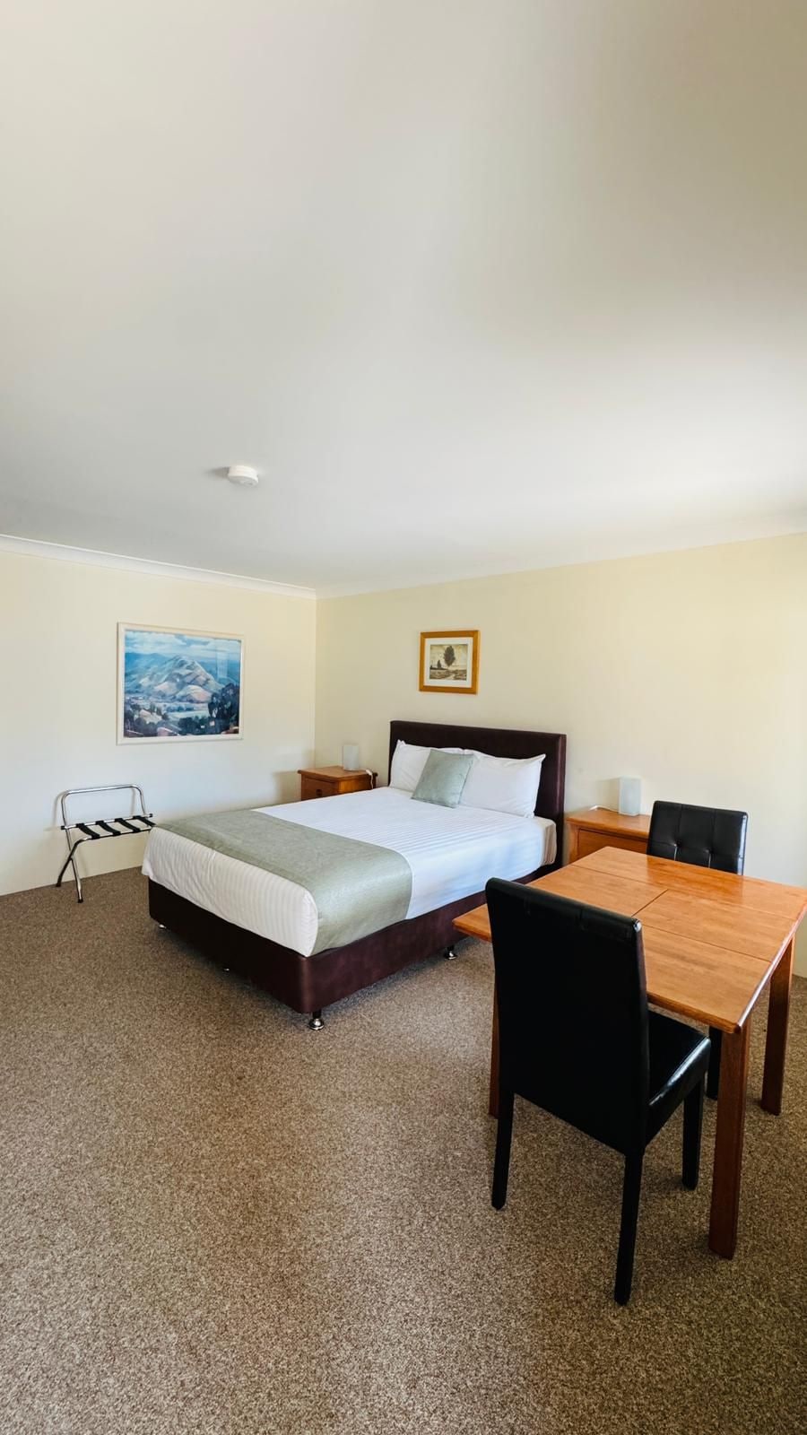 A hotel room with a bed, a table, and a chair. There is a painting on the wall and a picture frame.