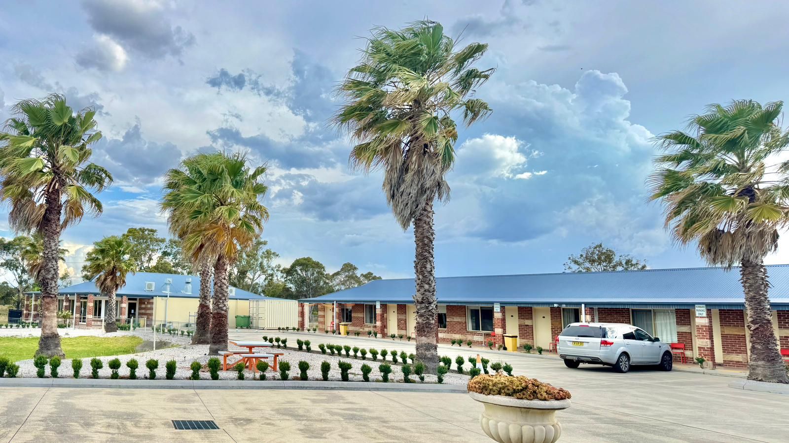 A hotel with a parking lot, palm trees, and a building with a blue roof.