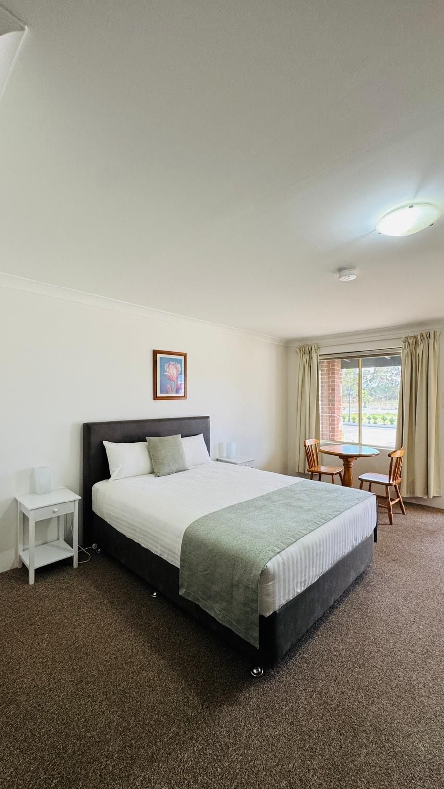 A hotel room with a large bed, a bedside table, a small table with chairs, and a window with curtains.