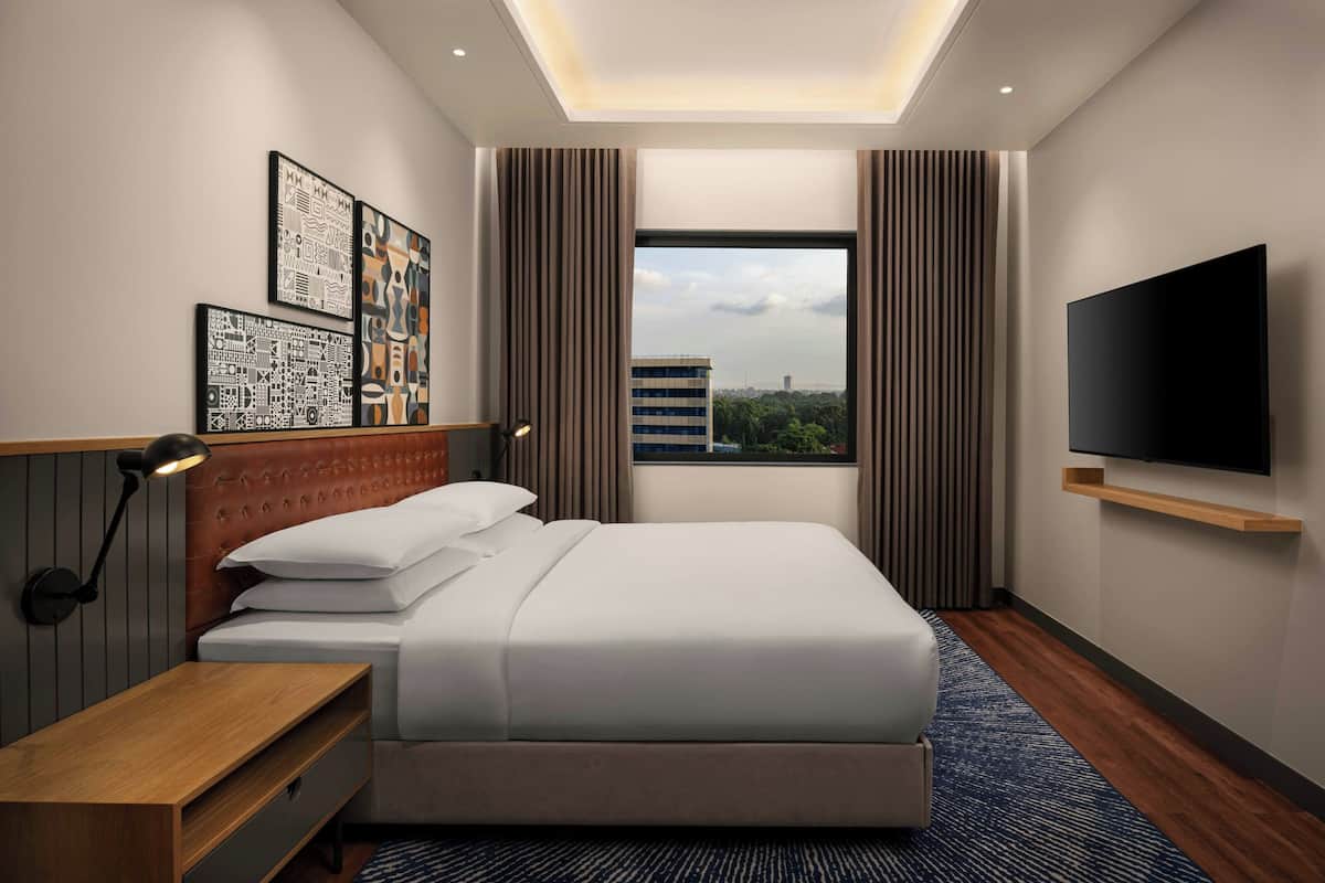 A modern hotel room with a large bed, a flat-screen TV mounted on the wall, a bedside table with a lamp, and artwork on the wall.