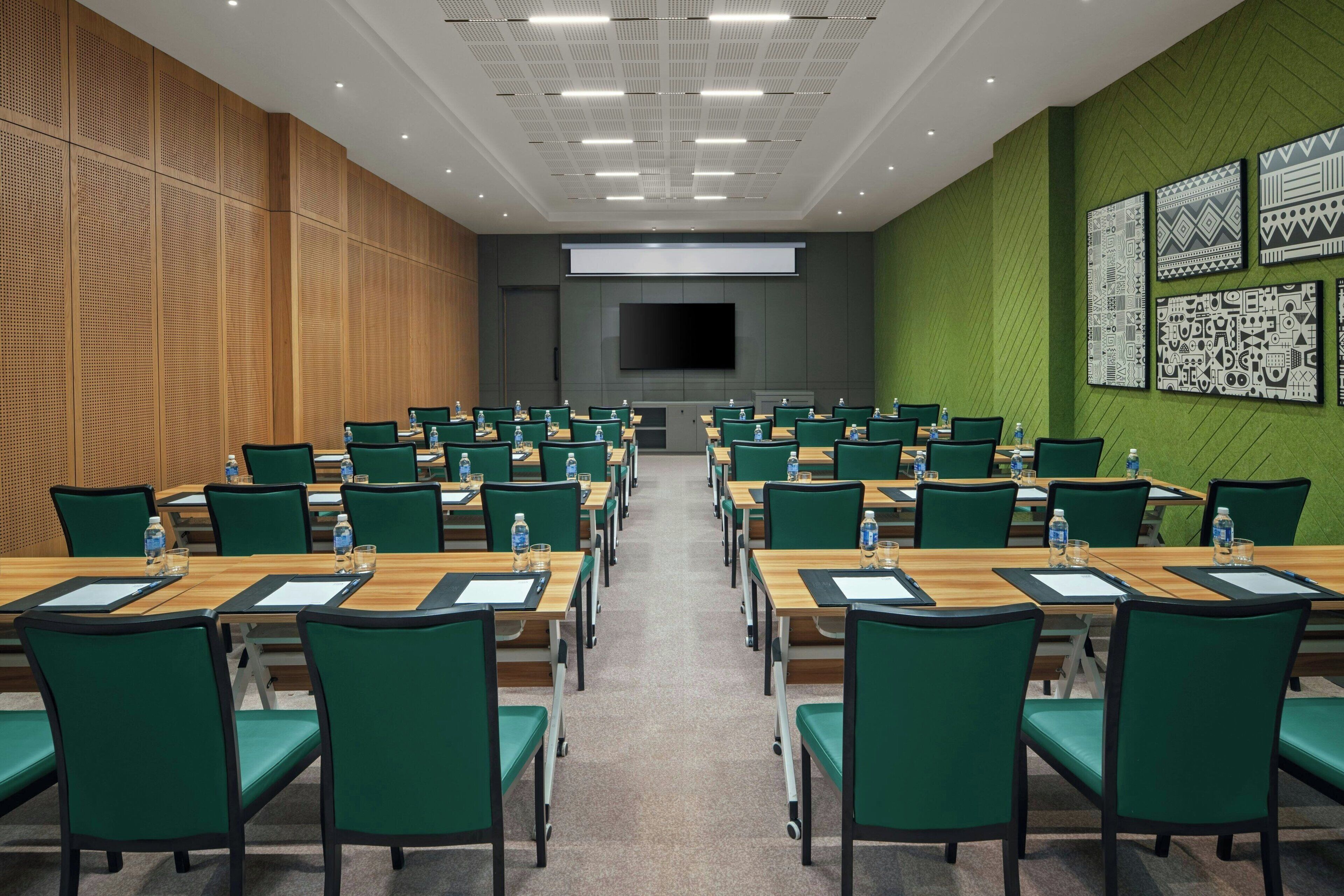 A conference room with rows of green chairs and wooden tables, a large screen, and patterned wall art.