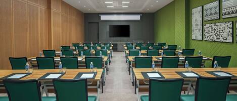 A conference room with rows of green chairs and wooden tables, a large screen, and patterned wall art.