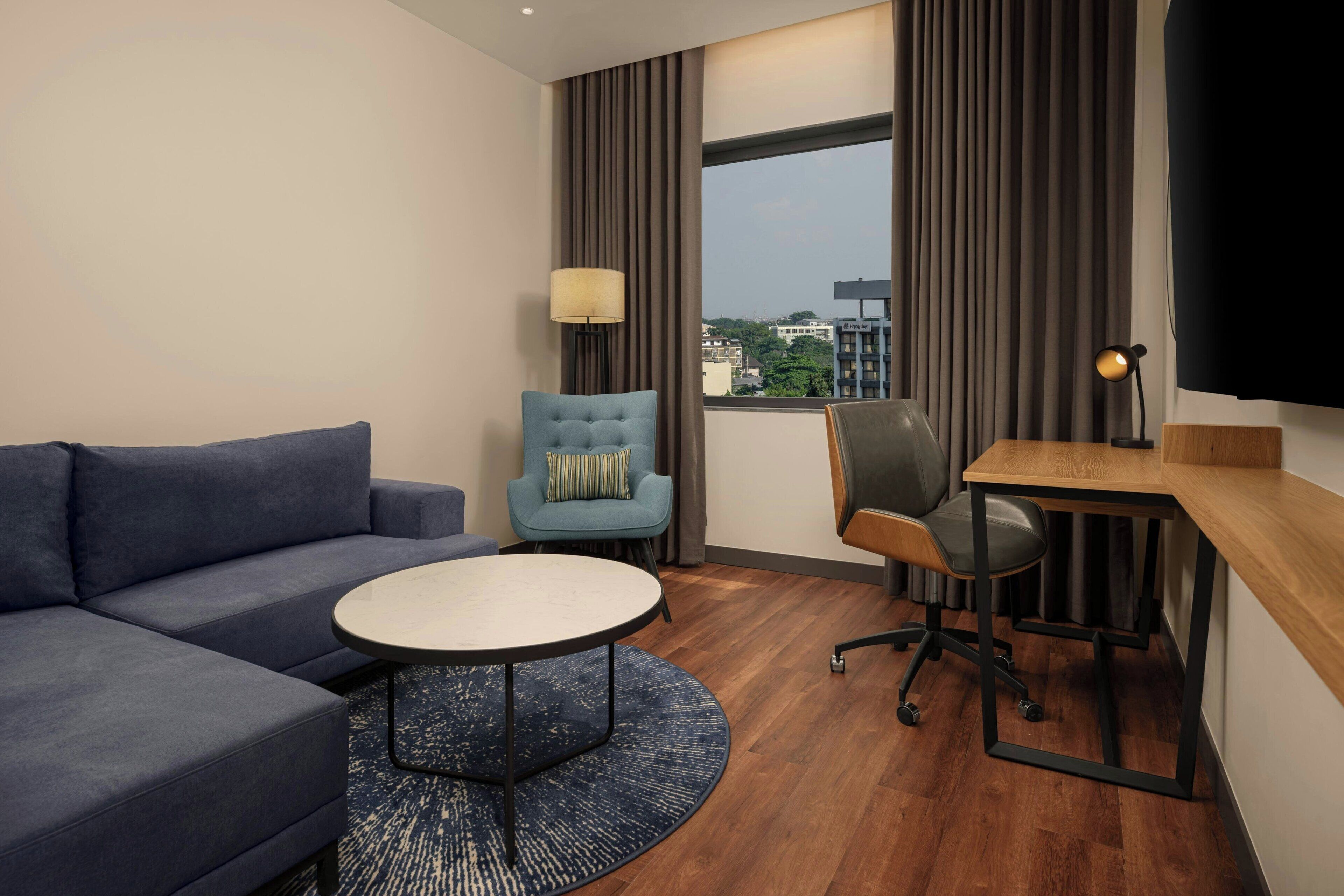 Une chambre d’hôtel moderne, équipée d’un canapé, d’une table basse ronde, d’un bureau avec une chaise, d’une lampe et d’un téléviseur.