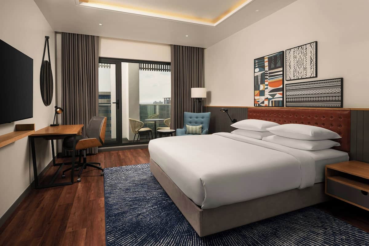 A modern hotel room with a large bed, a desk, a chair, a TV, and artwork on the wall.
