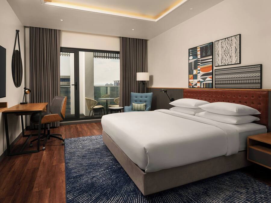 A modern hotel room with a large bed, a desk, a chair, a TV, and artwork on the wall.