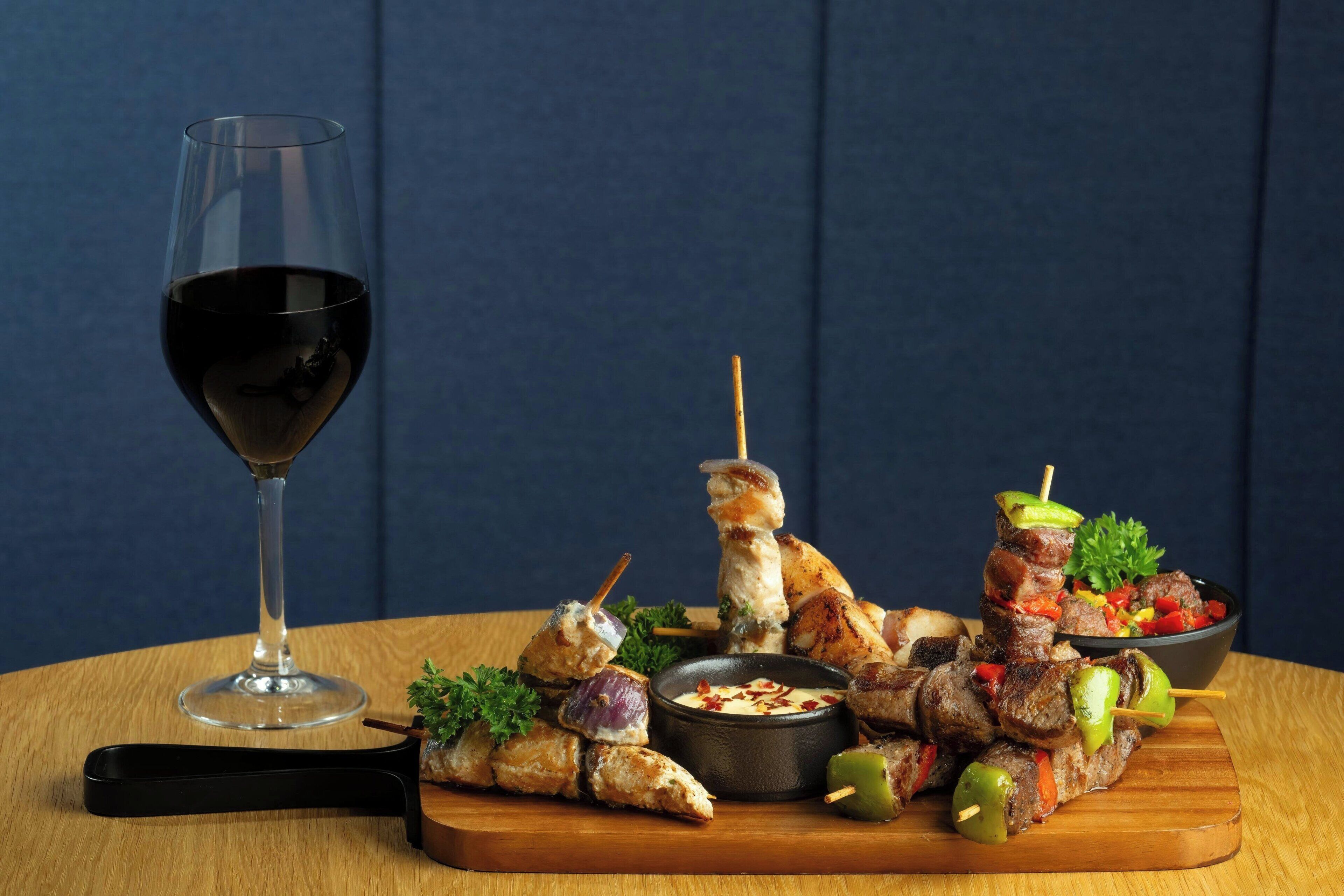 Ett träbord med spett av kött och grönsaker, en skål med dippsås och ett glas rött vin.