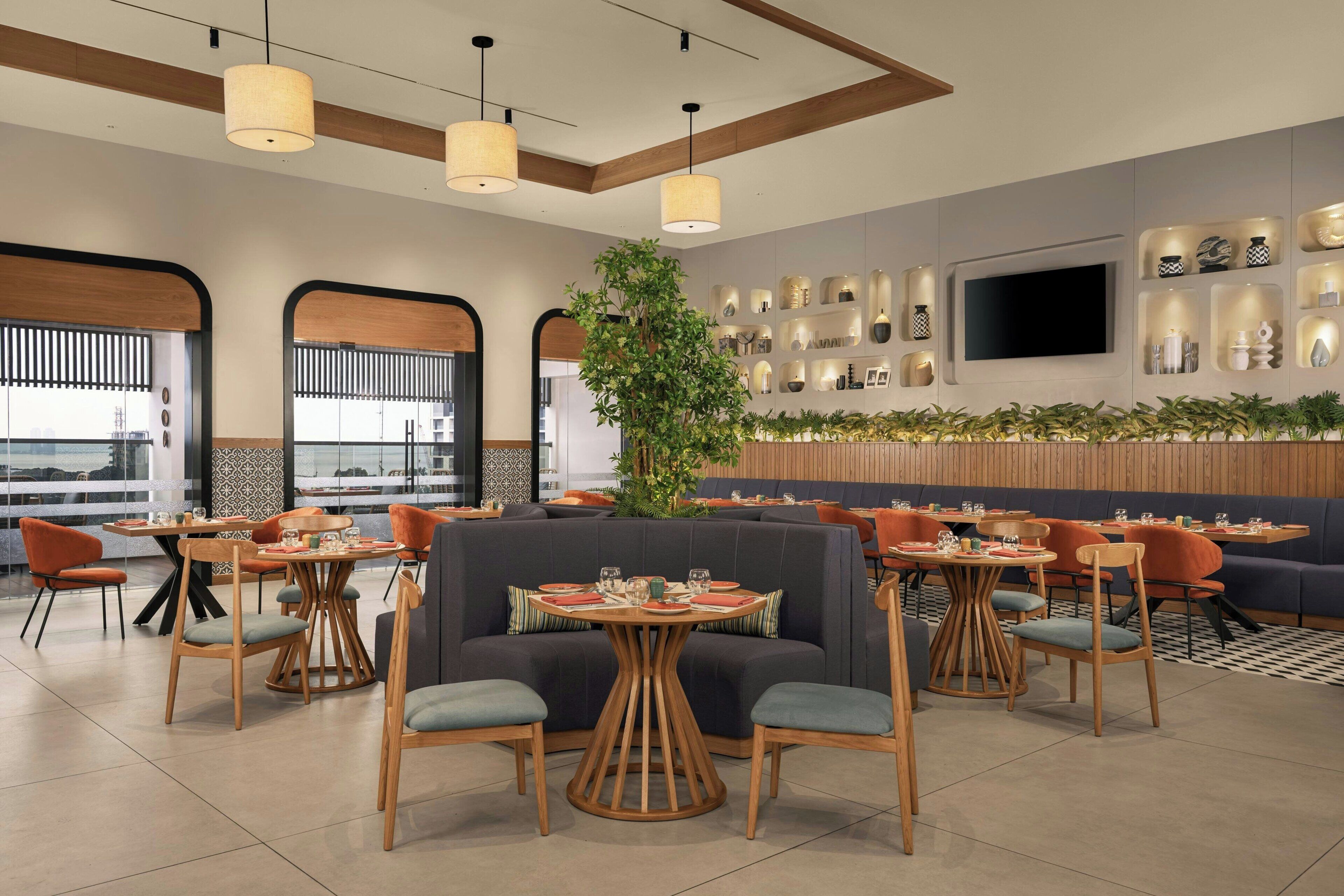 Un ristorante moderno con tavoli rotondi, sedute imbottite, illuminazione a sospensione, una pianta grande e una TV a parete.