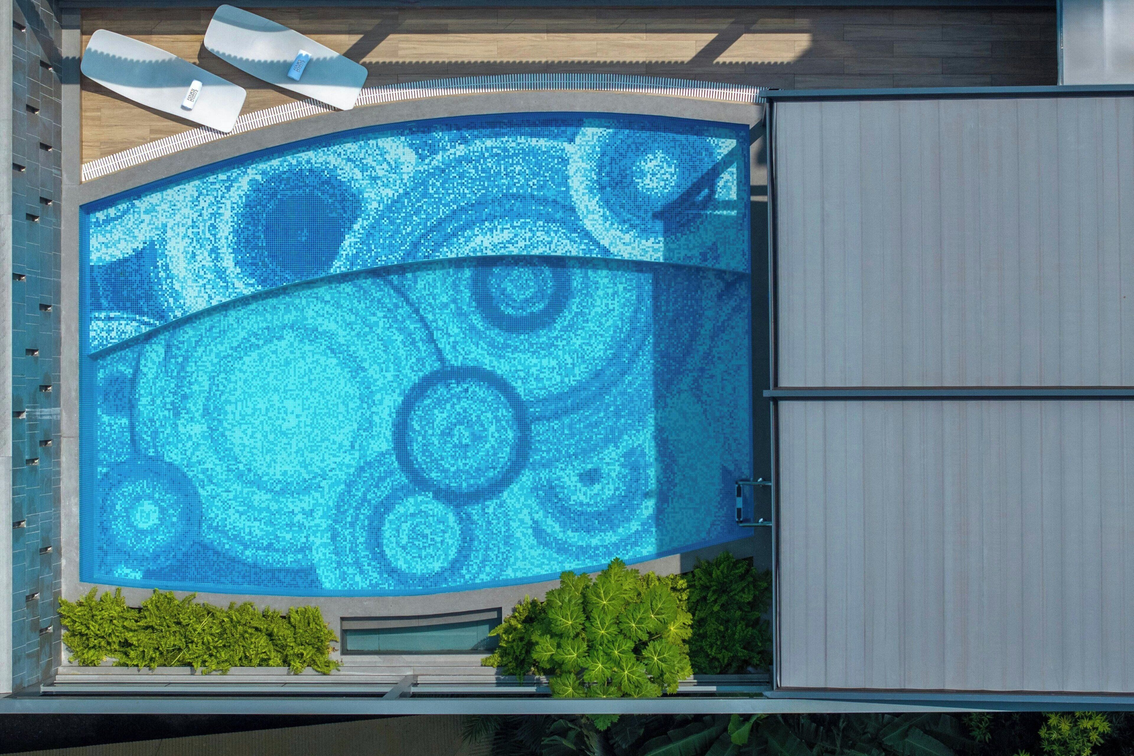 A modern building with a curved glass facade, a pool with a blue mosaic design, and a gated entrance.