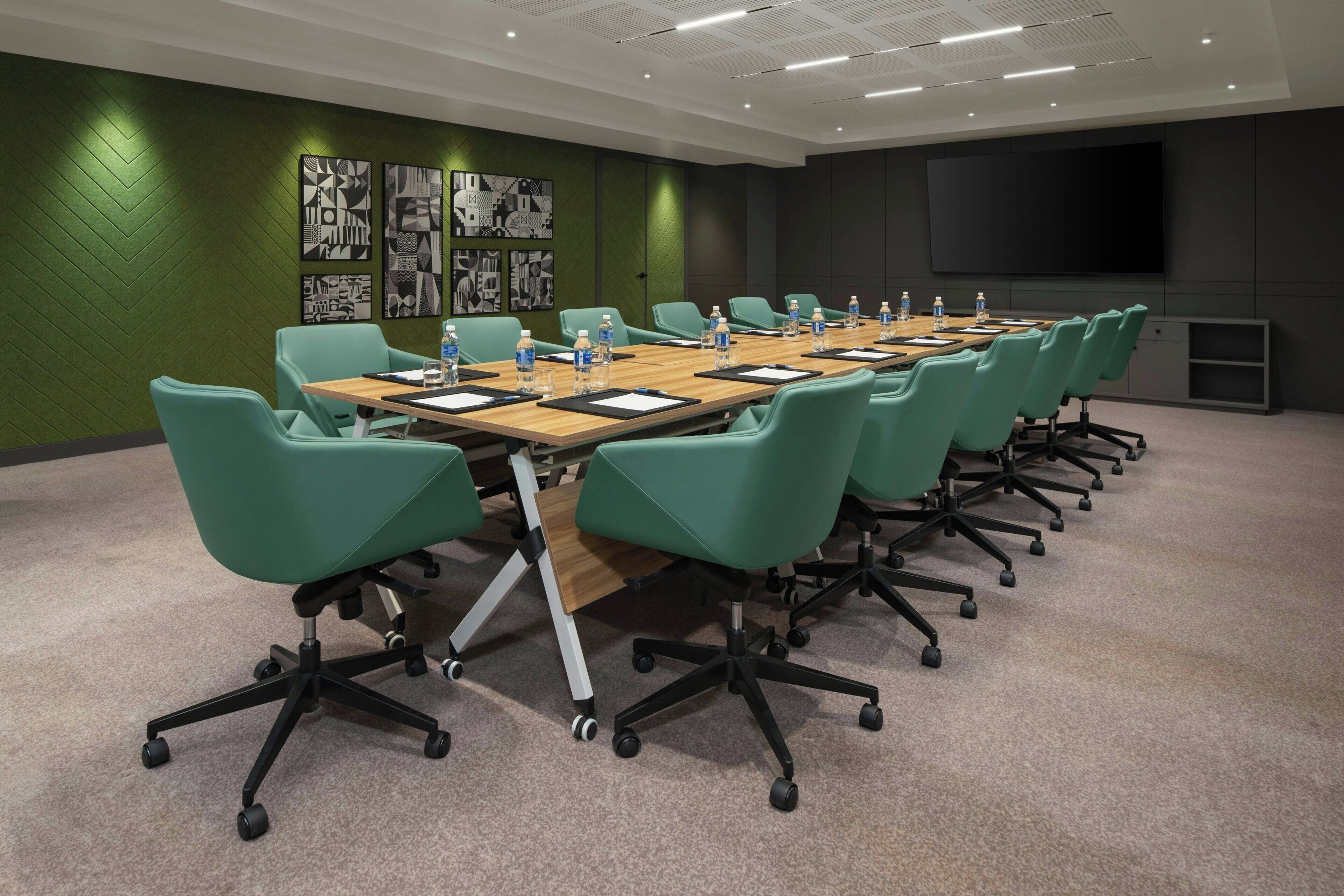 A conference room with a long table, green chairs, water bottles, and a large screen on the wall.