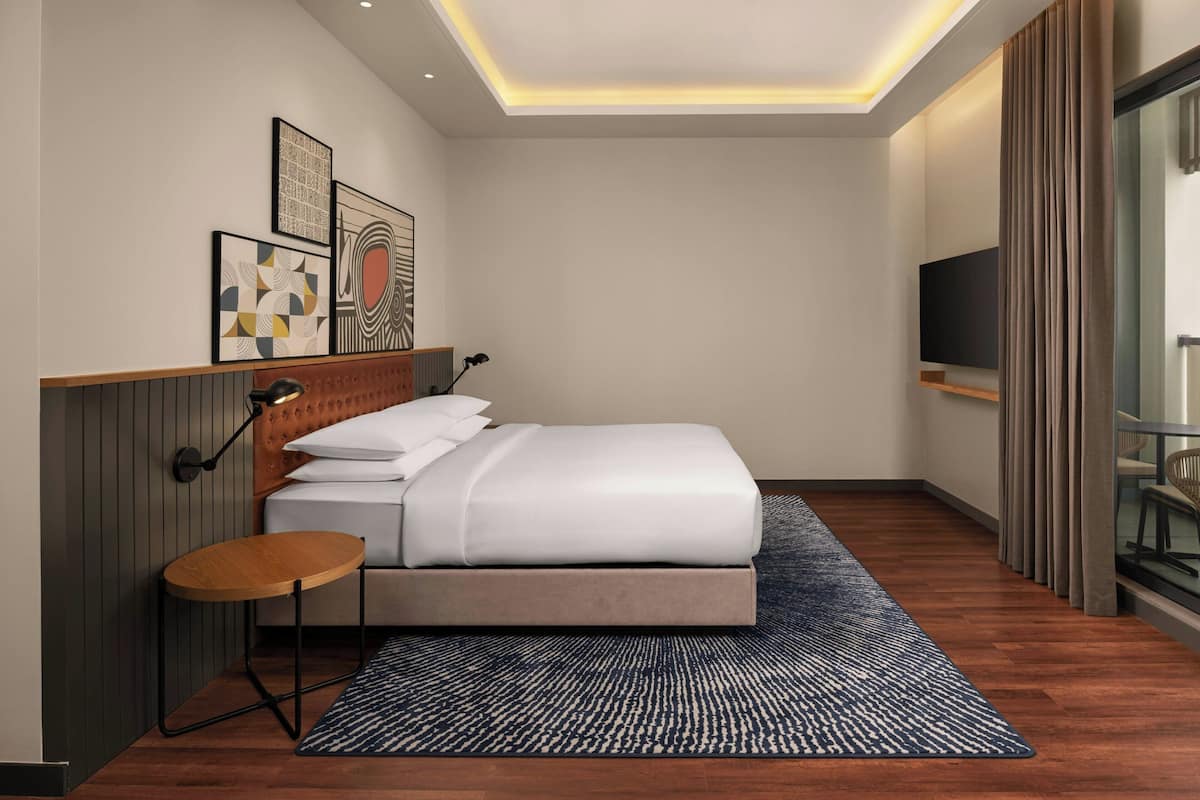 A modern hotel room with a neatly made bed, a small round table, a wall-mounted TV, and artwork on the wall.