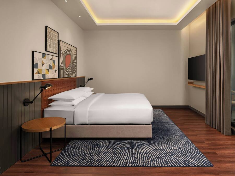 Een moderne hotelkamer met een netjes opgemaakt bed, een klein rond tafeltje, een wandtelevisie en kunst aan de muur.
