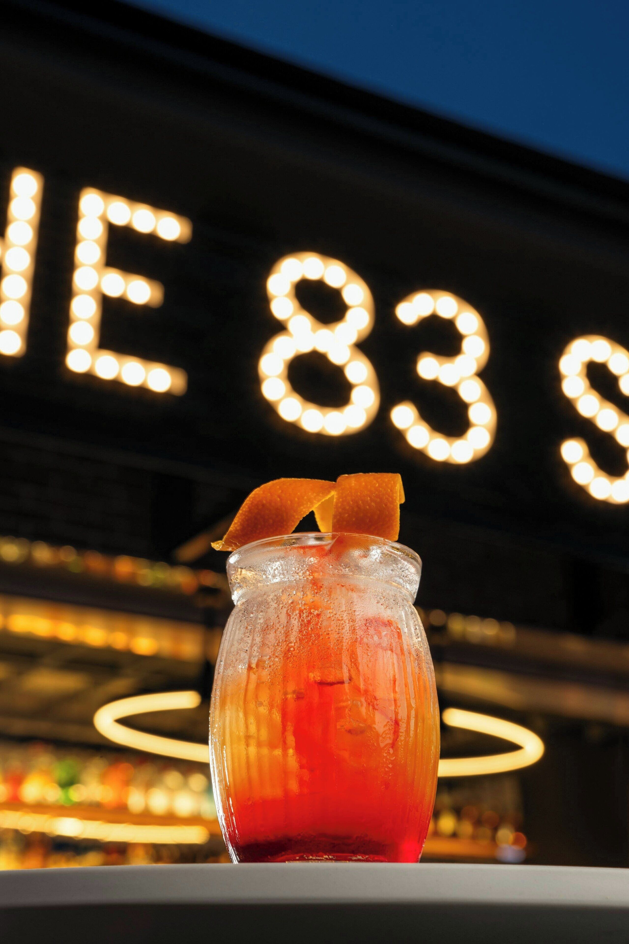 Un cocktail servi dans un verre, agrémenté d’une garniture, présenté sur fond de lettres illuminées et d’un comptoir de bar.