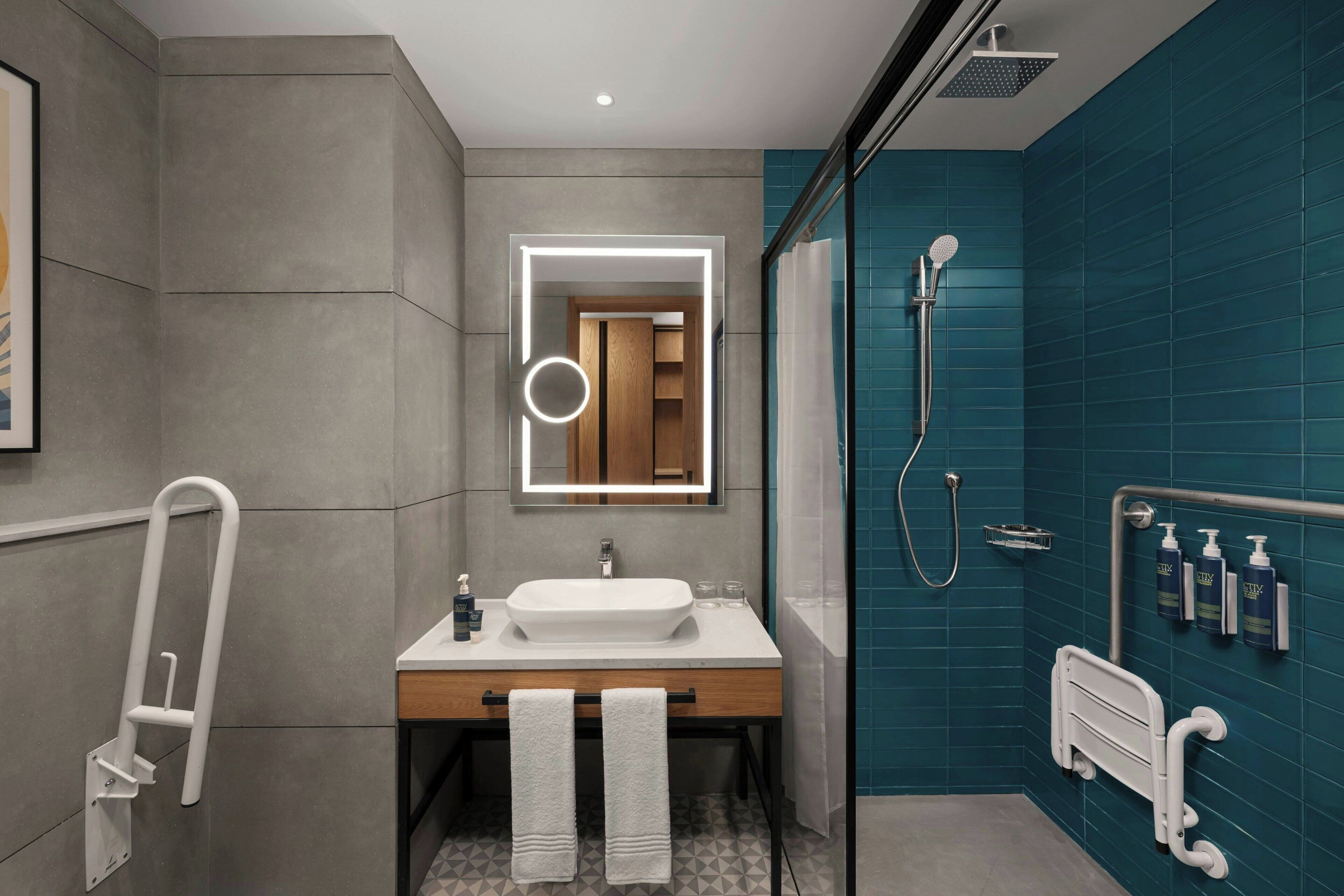 A modern bathroom with a white sink, a mirror with lights, a shower with a handheld showerhead, and a grab bar.