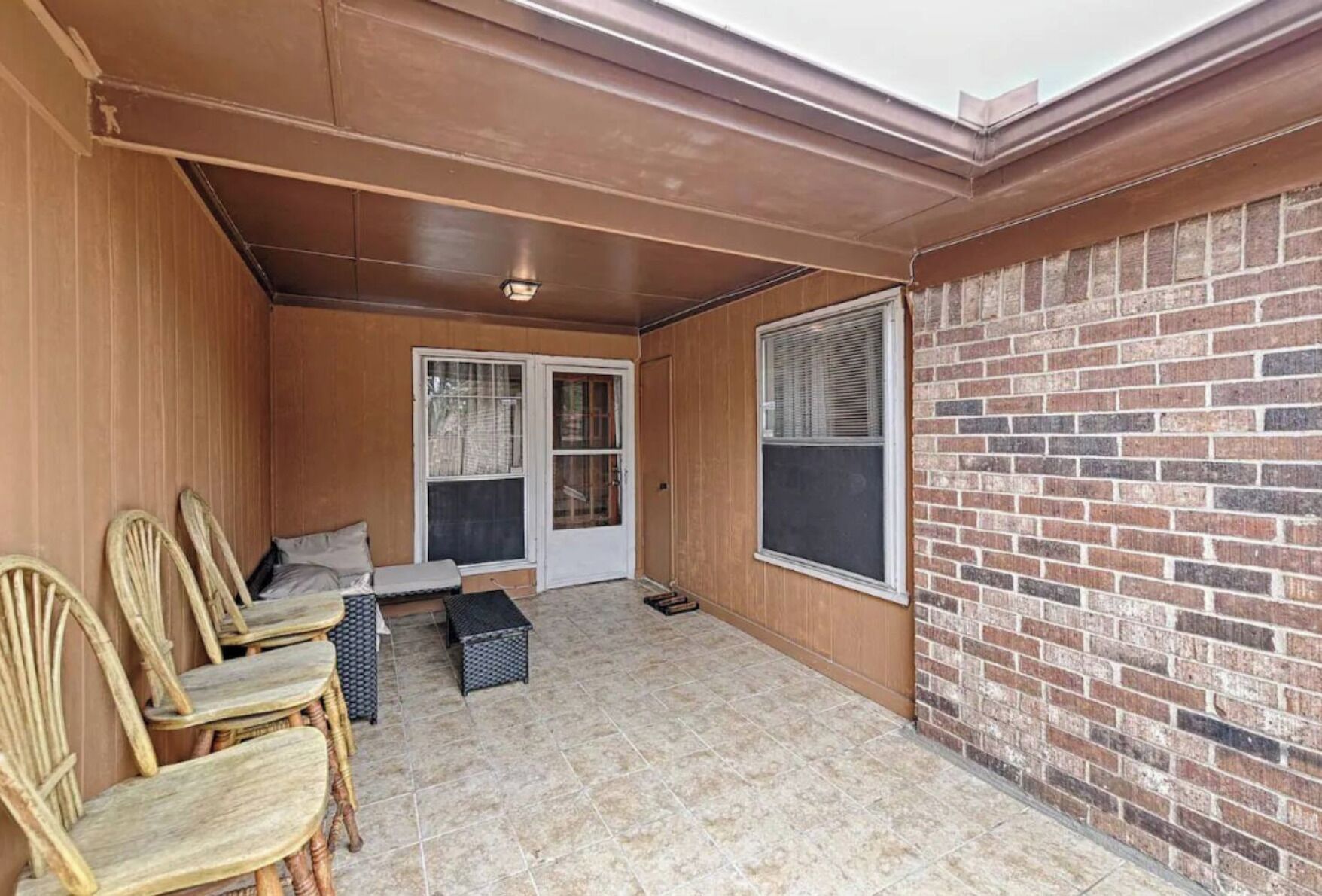 A covered patio with a brick wall, a sliding glass door, and a seating area with chairs and a small table.