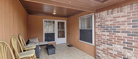 A covered patio with a brick wall, a sliding glass door, and a seating area with chairs and a small table.