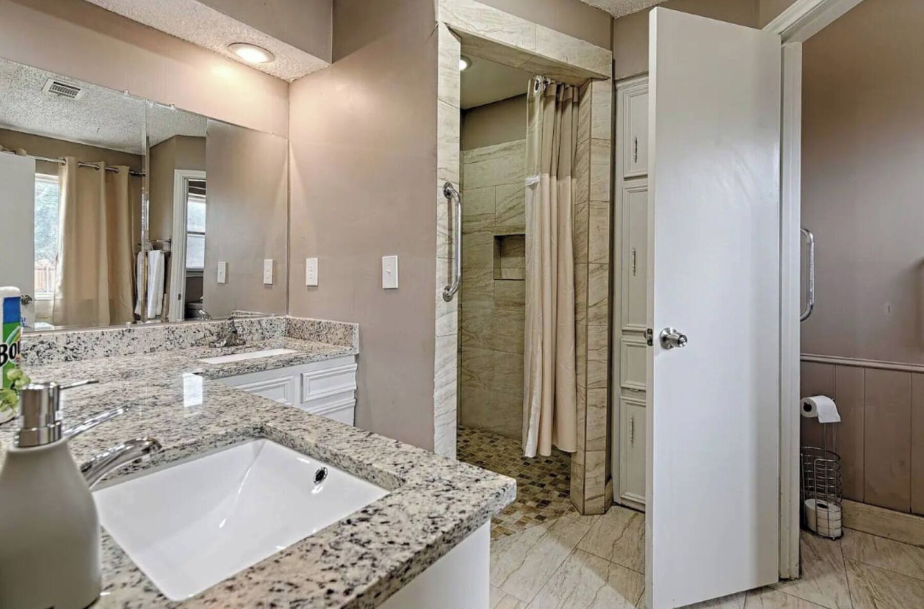 Un baño con lavamanos de encimera de granito, ducha con puerta de cristal y espejo sobre el lavamanos.