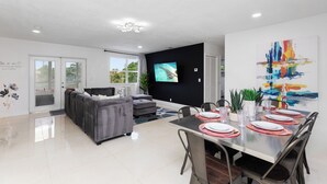 A modern living room with a grey sofa, a dining table set for four, and a flat-screen TV mounted on the wall. - Luxe Coastal Escape King BD Heated pool WaterFront (Pompano Beach)
