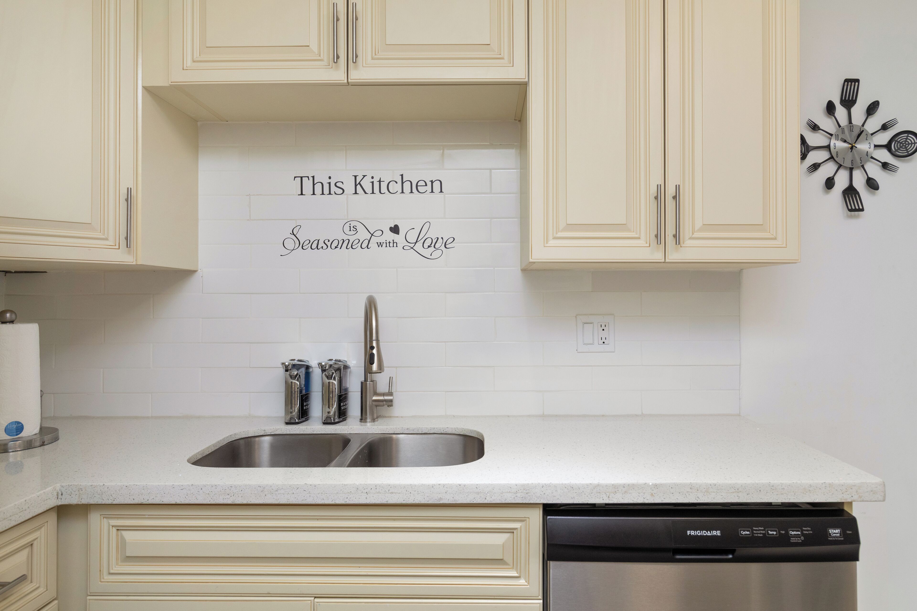 A kitchen with white cabinets, a stainless steel sink, and a dishwasher. A decorative sign on the wall reads 'This Kitchen is Seasoned with Love.'