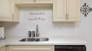 A kitchen with white cabinets, a stainless steel sink, and a dishwasher. A decorative sign on the wall reads 'This Kitchen is Seasoned with Love.'