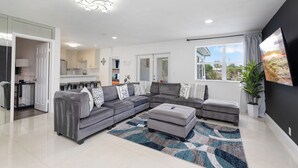 A modern living room with a grey sectional sofa, a matching ottoman, and a patterned rug. There is a dining area with a table and chairs, and a kitchen visible through an open door. - Luxe Coastal Escape King BD Heated pool WaterFront (Pompano Beach)
