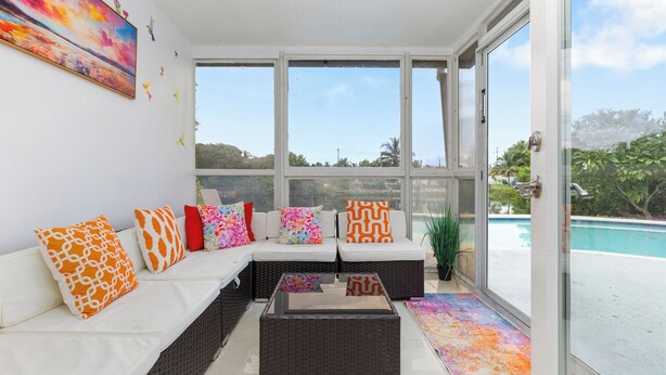 A bright, sunlit room with a white sofa, colorful cushions, a glass coffee table, and a view of a swimming pool. - Luxe Coastal Escape King BD Heated pool WaterFront (Pompano Beach)