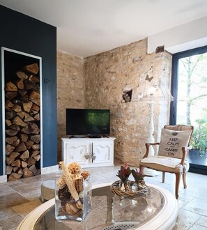 Una sala de estar con una pared de piedra, un televisor, una mesa de centro de vidrio con adornos y una silla con cojín, además de una pila de leña.