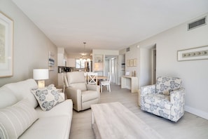 Un salon spacieux avec un canapé, un fauteuil et une table basse. On aperçoit à l’arrière-plan un coin repas avec une table et des chaises.