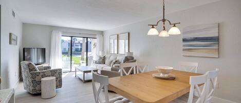 A spacious living area with a wooden dining table, white chairs, a sofa, and a television.